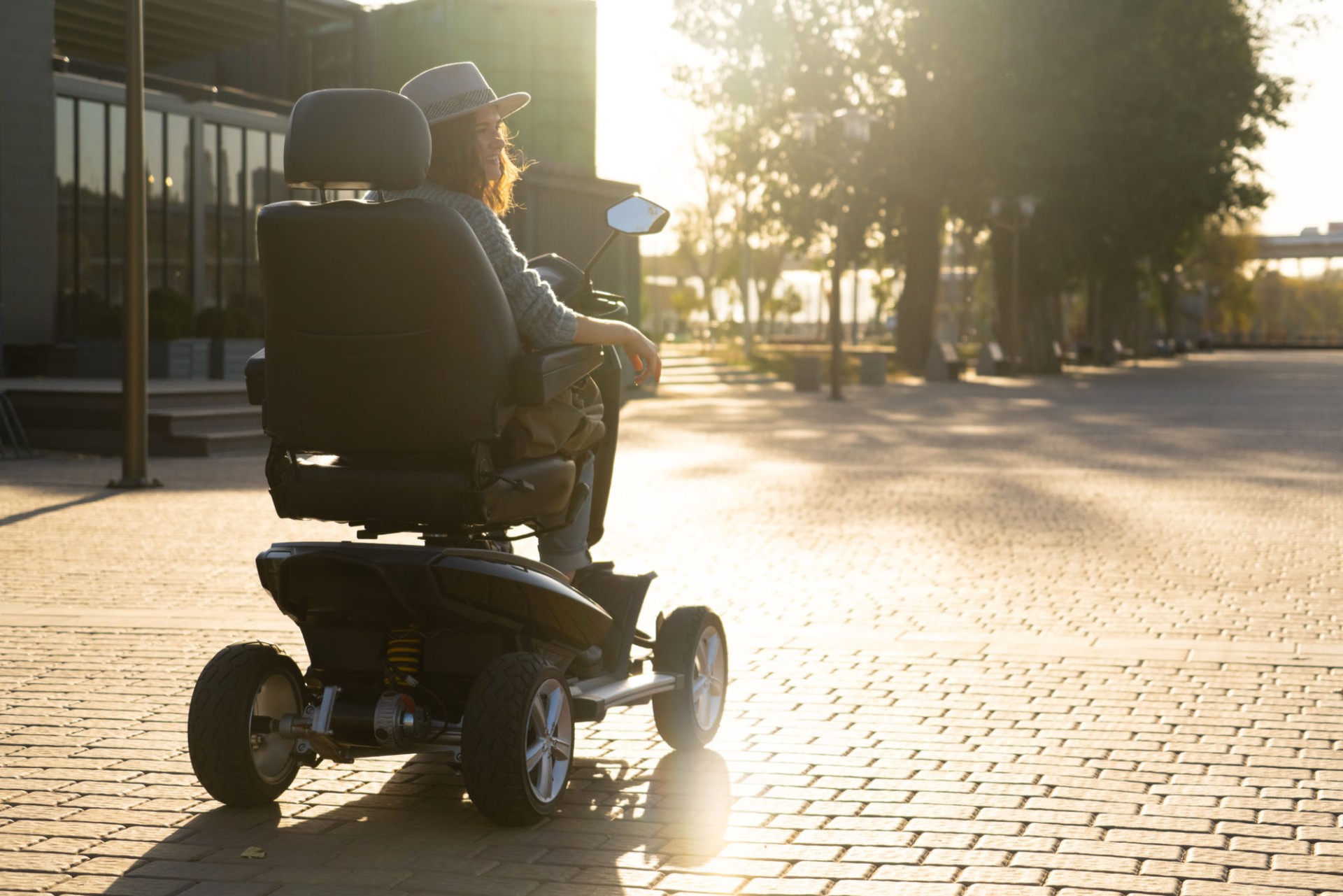 Woman tourist wearing gray hat and sweater is riding a four wheel mobility electric scooter on a city street. Sunset back light.