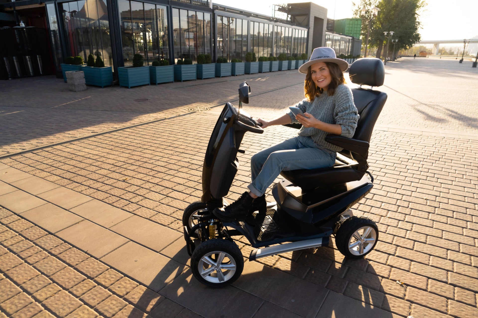 Woman tourist wearing gray hat and sweater is riding a four wheel mobility electric scooter on a city street. Sunset back light.